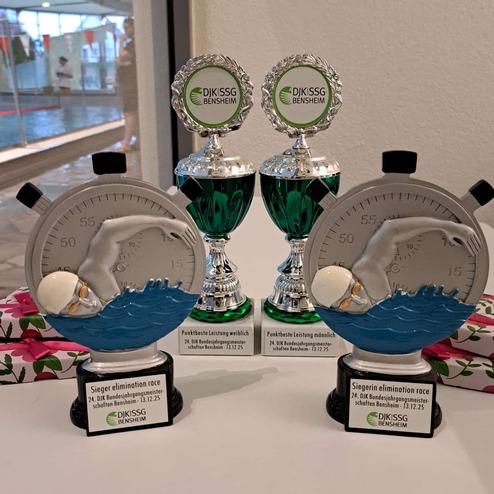 Drei Pokale und ein Zeitmesser stehen auf einem Tisch, umgeben von bunten Geschenkboxen im Hintergrund.