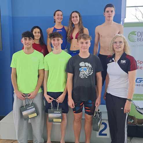 Gruppenfoto von sechs Schwimmern und einer Trainerin auf einem Podium nach einem Wettkampf, alle l&auml;cheln gl&uuml;cklich.
