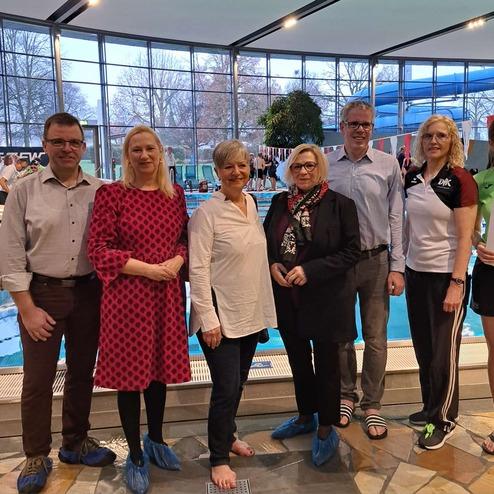 Gruppe von sechs Personen in Schwimmbad, eine Person h&auml;lt ein Dokument, alle tragen Badeschuhe.