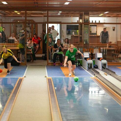 Zwei Spielerinnen werfen Bowlingkugeln auf einer Bahn, Zuschauer sitzen in der Hintergrund, Innenszene einer Kegelbahn.