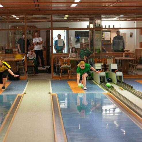 Zwei Spieler in Sportkleidung werfen Bowlingkugeln auf zwei Bahnen in einem gut besuchten Bowlingcenter.