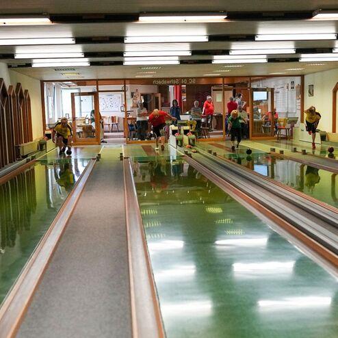 Innenansicht einer Kegelbahn mit zwei parallelen Bahnen und Personen, die kegeln und zuschauen. Holzverkleidung an den W&auml;nden.