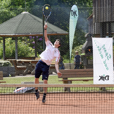 Ein Tennisspieler springt, um einen Ball &uuml;ber das Netz auf einem roten Sandplatz zu schlagen. Im Hintergrund sind B&auml;ume.