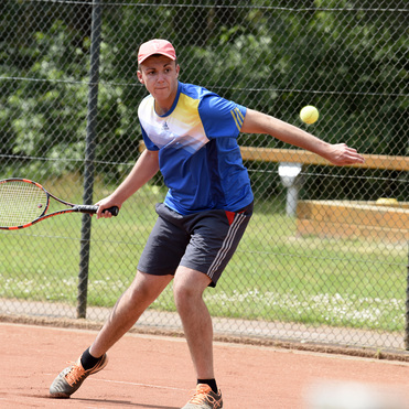 Junger Mann in blauer Sportbekleidung spielt Tennis auf einem Sandplatz, w&auml;hrend er einen Ball mit dem Schl&auml;ger trifft.
