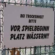 Hinweis-Schild an einem Zaun: "Bei Trockenheit bitte vor Spielbeginn Platz w&auml;ssern!" in klaren Buchstaben.