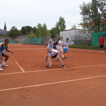 Gruppe von f&uuml;nf Kindern beim Tennistraining auf einem Sandplatz, umgeben von B&auml;umen und einem Geb&auml;ude im Hintergrund.