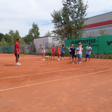 Gruppenunterricht auf einem Tennisplatz, Trainer zeigt Technik f&uuml;r Sch&uuml;lerinnen und Sch&uuml;ler, die Tennisb&auml;lle halten.