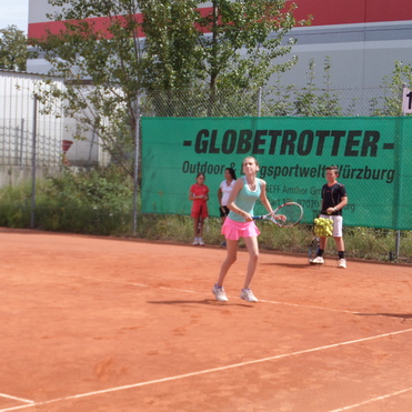 M&auml;dchen mit Tennisracket steht auf einem Sandplatz, w&auml;hrend Kinder im Hintergrund zuschauen. Gr&uuml;ne Werbebande sichtbar.