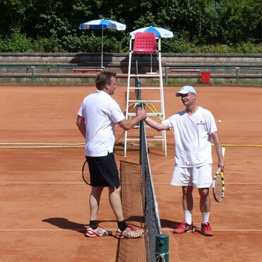 Zwei Tennisspieler stehen am Netz und unterhalten sich auf einem roten Sandplatz. Im Hintergrund steht ein Schiedsrichterstuhl.