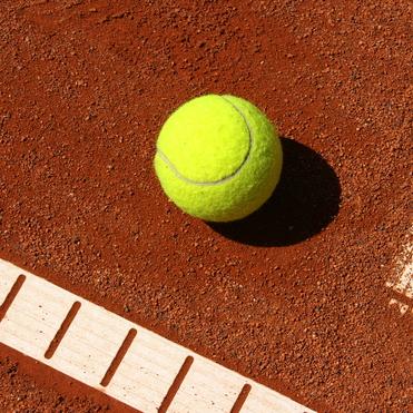 Eine leuchtend gelbe Tennisball liegt auf einem roten Sandplatz, nahe den wei&szlig;en Markierungen.