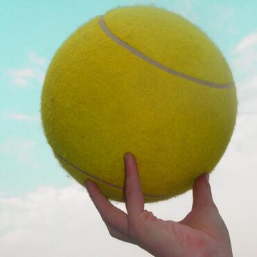 Eine Hand h&auml;lt einen gro&szlig;en, gelben Tennisball vor einem hellblauen Himmel mit wenigen Wolken.