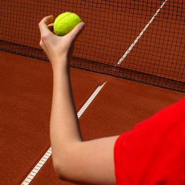 Nahaufnahme einer Hand, die einen Tennisball &uuml;ber einer roten Sandplatz-Tennislinie h&auml;lt, mit Netz im Hintergrund.