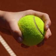 Nahaufnahme einer Hand, die einen gelben Tennisball festh&auml;lt, vor einem roten Tennisplatz.