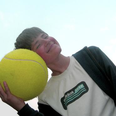 Junger Mann mit einem gro&szlig;en Tennisball, l&auml;chelnd und mit dem Ball an seinen Kopf gelehnt, vor einem blauen Himmel.