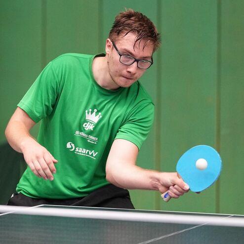 Junger Mann in gr&uuml;nem T-Shirt spielt Tischtennis, h&auml;lt einen Schl&auml;ger und zielt auf einen Ball an der Platte.