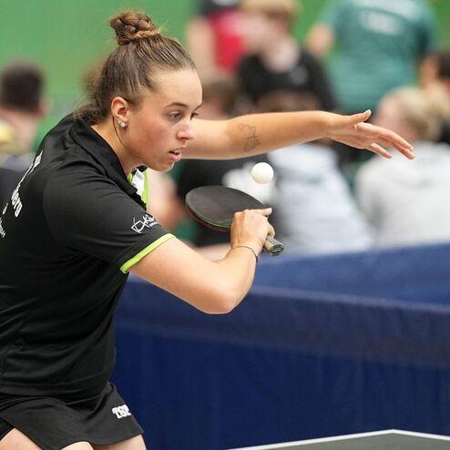 Junge Tischtennisspielerin bereitet einen Schlag vor, Hintergrund mit Zuschauern und Tischtennisplatten sichtbar.