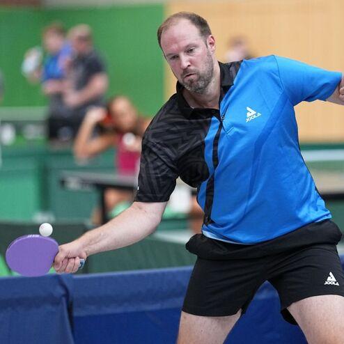 Ein Spieler schl&auml;gt einen Tischtennisball mit einem lila Schl&auml;ger in einem Hallenspielbereich.