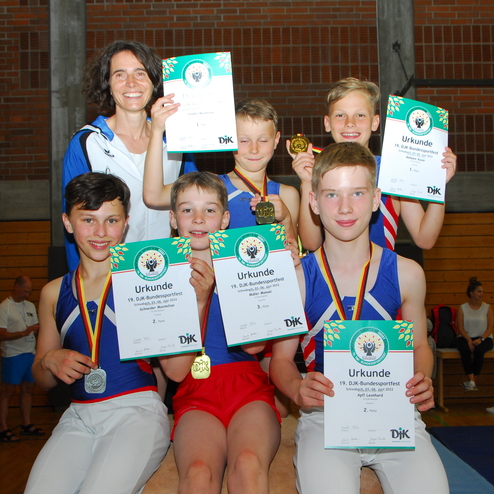 Sechs Kinder mit Medaillen und Urkunden posieren l&auml;chelnd mit einer Trainerin in einer Sporthalle.