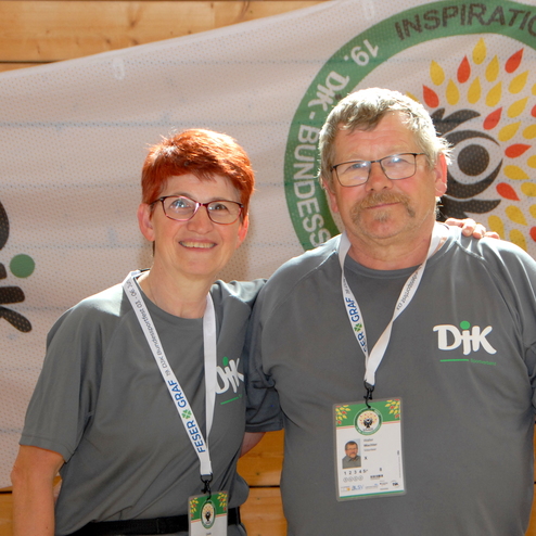 Paar mit grauen T-Shirts steht vor einer gro&szlig;en, bemalten Wand mit Logo und Schriftzug der Veranstaltung.