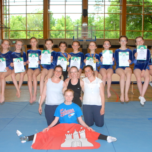 Gruppennfoto von Turnerinnen mit Medaillen und Urkunden auf dem Schwebebalken in einer Turnhalle.