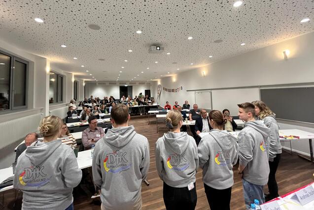 Sechs Personen in grauen Hoodies stehen vor einer Gruppe von Teilnehmern in einem modernen Seminarraum.