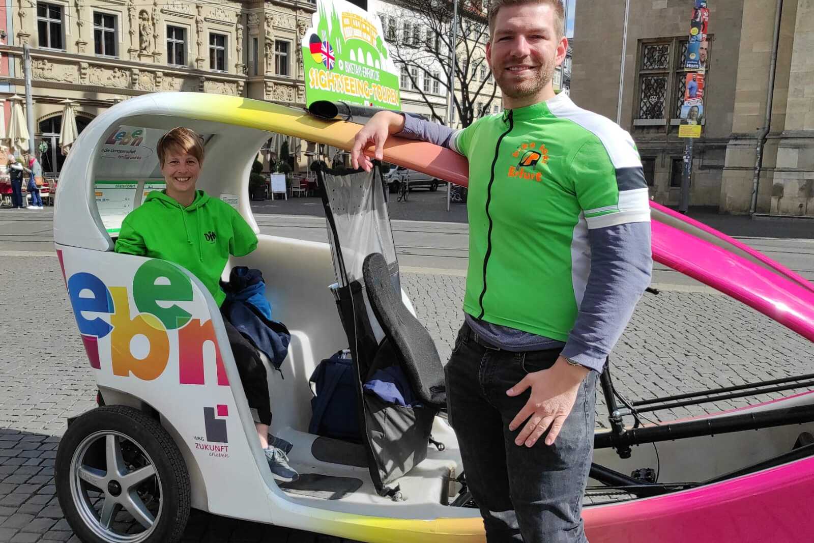 Zwei Personen in gr&uuml;nen Shirts stehen neben einem bunten Pedelac in einer Stadt, im Hintergrund historische Geb&auml;ude.