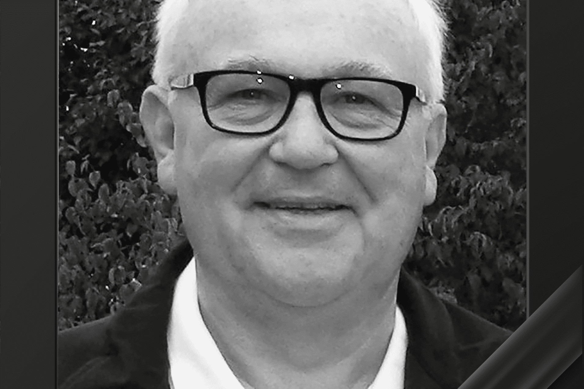 Schwarz-wei&szlig;e Portraitaufnahme eines &auml;lteren Mannes mit Brille, l&auml;chelnd vor einem gr&uuml;nen Hintergrund.