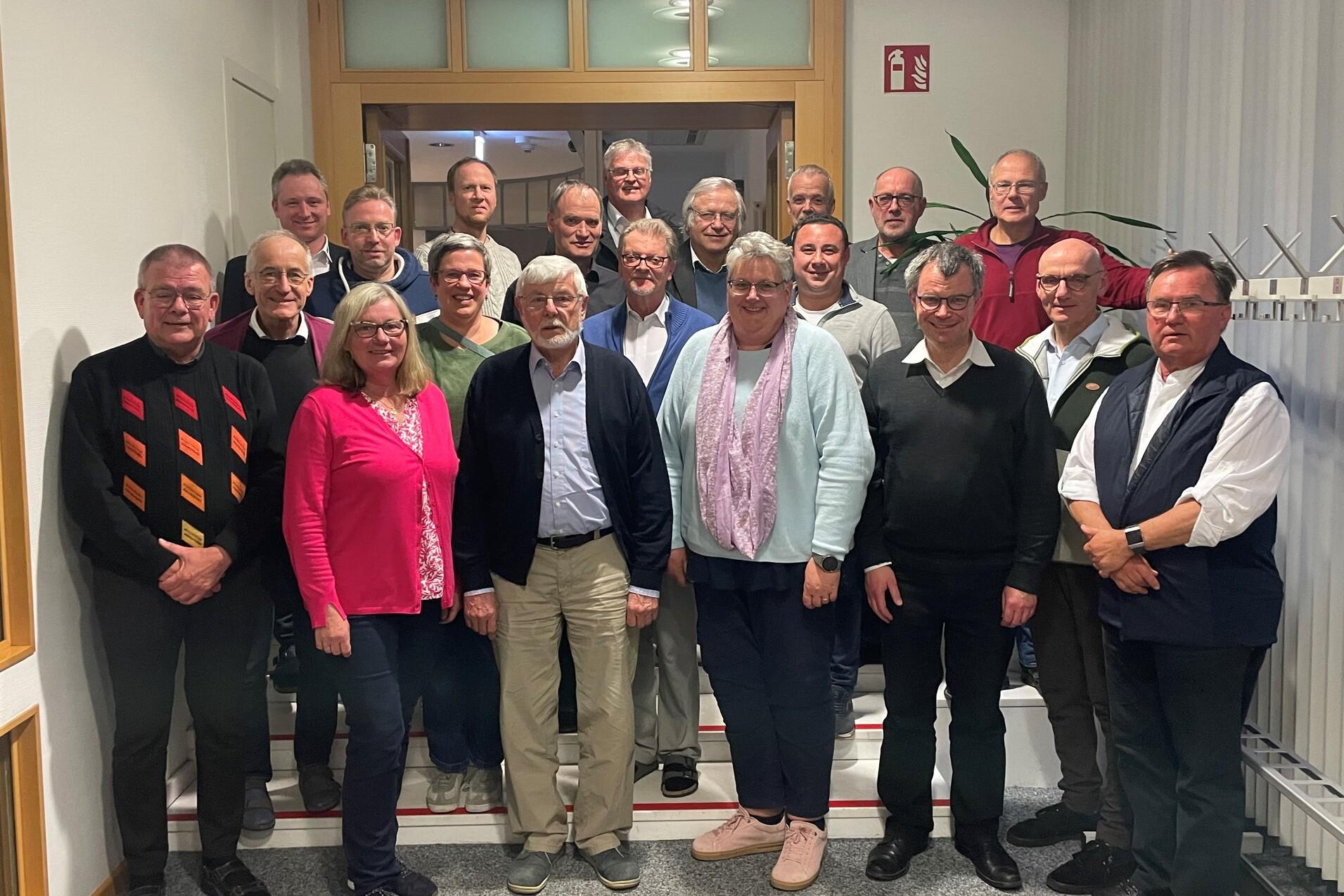 Gruppenfoto von 24 Personen in einem Flur, einige stehen, andere sitzen, alle blicken in die Kamera.