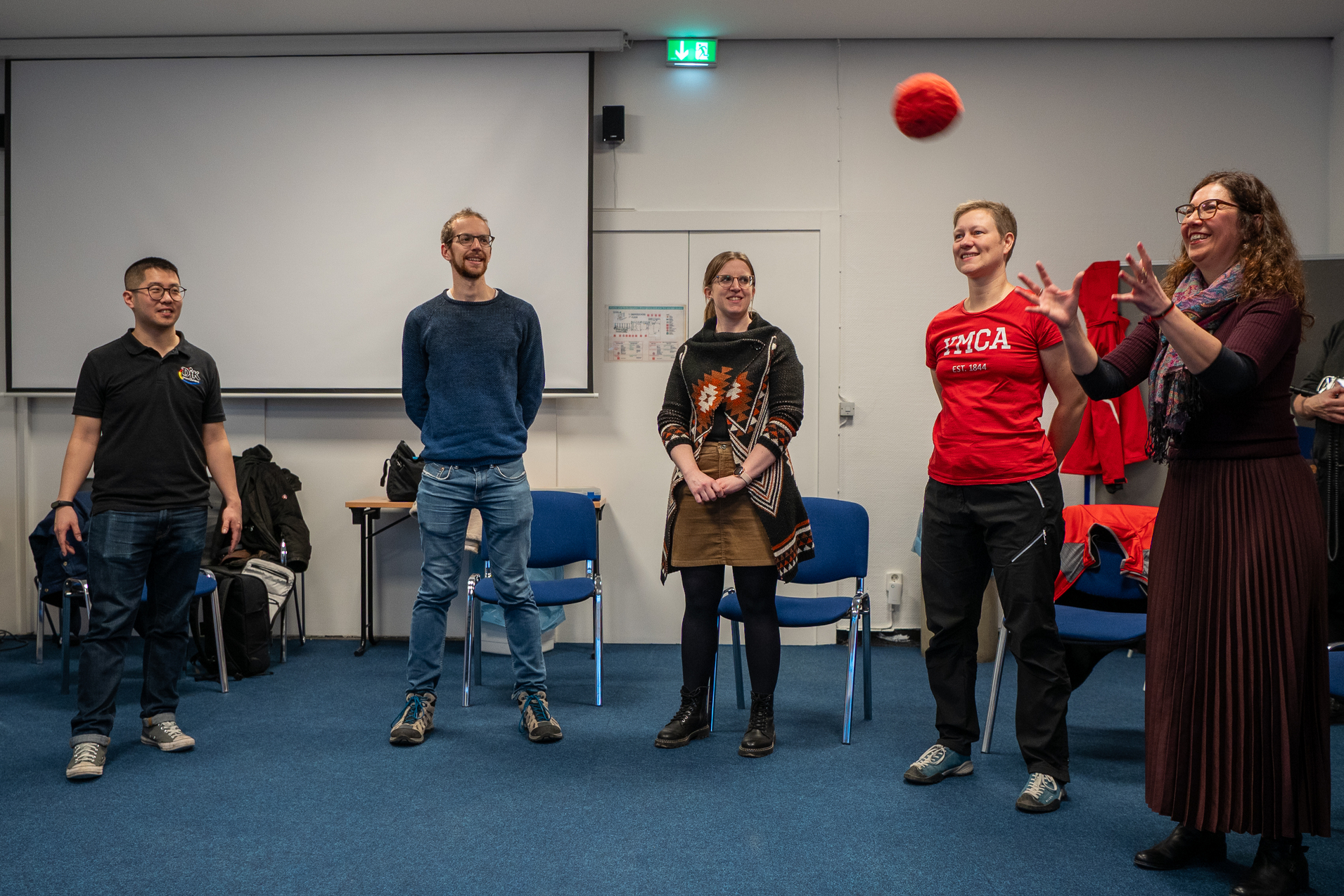 F&uuml;nf Personen stehen in einem Seminarraum und spielen mit einem roten Ball. Stuhlreihen im Hintergrund.