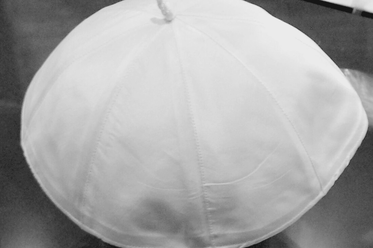 Wei&szlig;e Kappe aus Stoff, leicht gew&ouml;lbt mit einer kleinen Spitze oben, auf einem dunklen Untergrund.