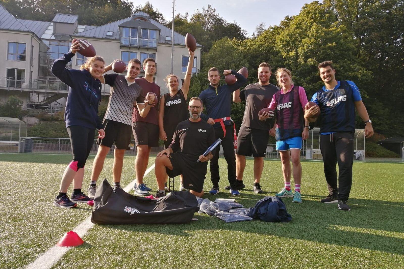 Eine Gruppe von neun Personen h&auml;lt American Footballs auf einem Sportplatz mit Kunstrasen. Im Hintergrund sind B&auml;ume und ein Geb&auml;ude.