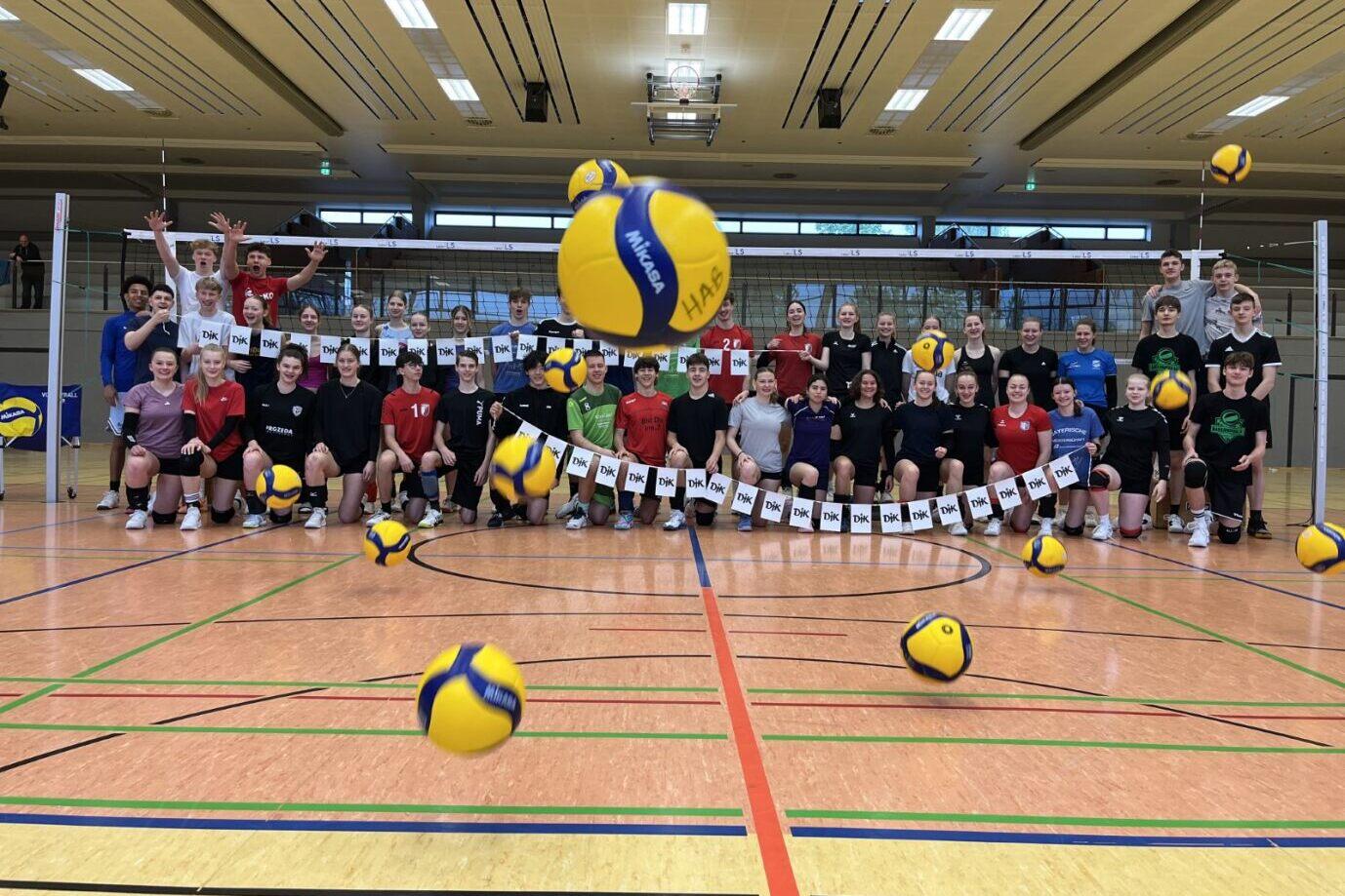 Gruppenfoto von Volleyballspielern in einer Sporthalle, mit mehreren B&auml;llen in der Luft und bunten Trikots.