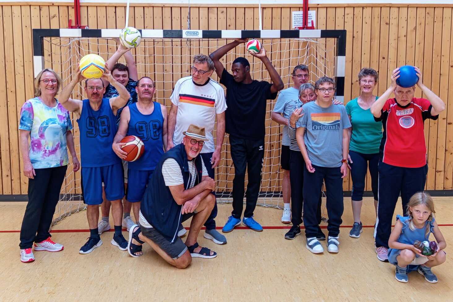 Gruppe von sportlichen Menschen in einer Sporthalle, die verschiedene B&auml;lle vor einem Tor halten und l&auml;cheln.