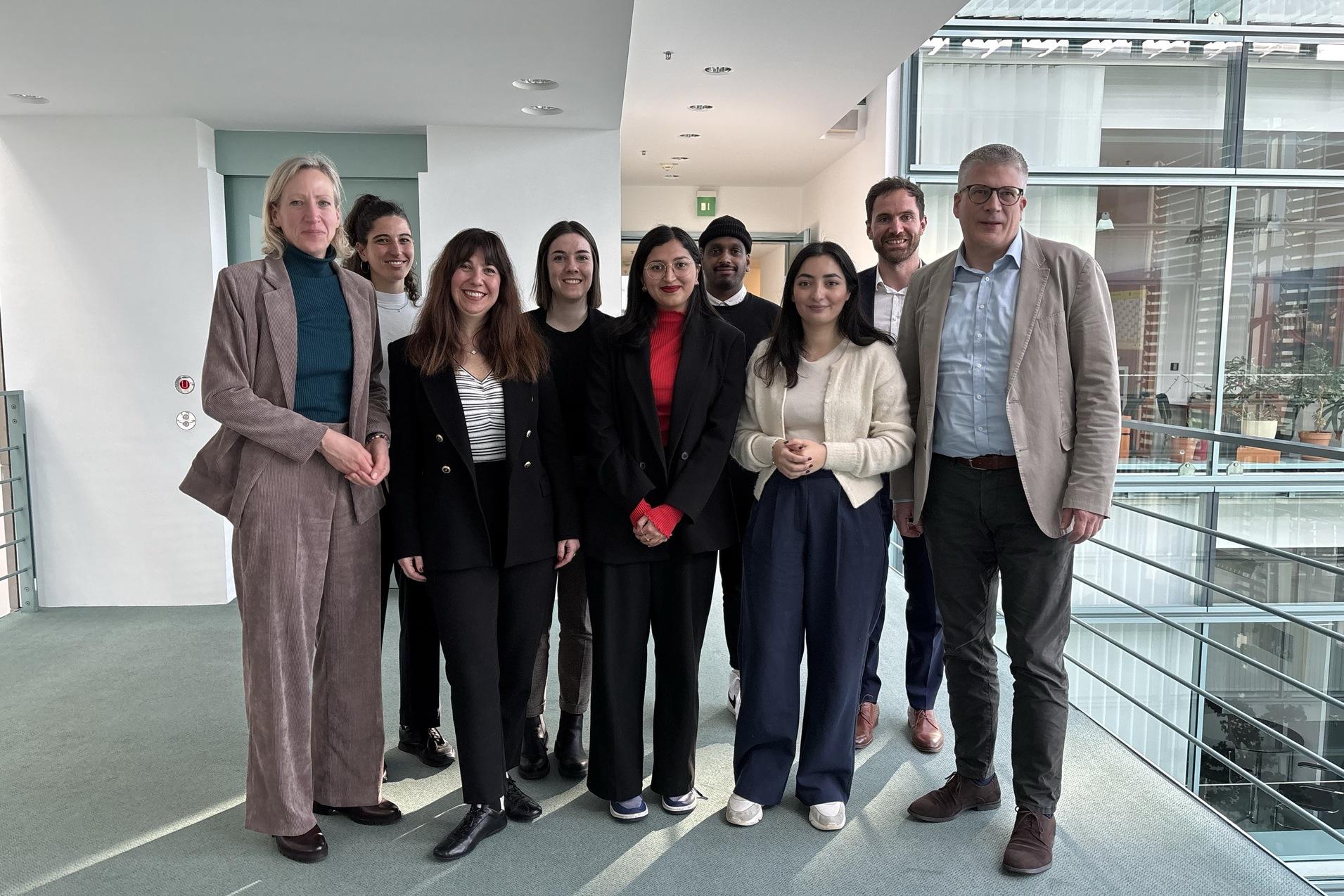 Gruppennfoto von acht Personen in Businesskleidung, stehend in einem modernen Innenraum mit gro&szlig;en Fenstern.