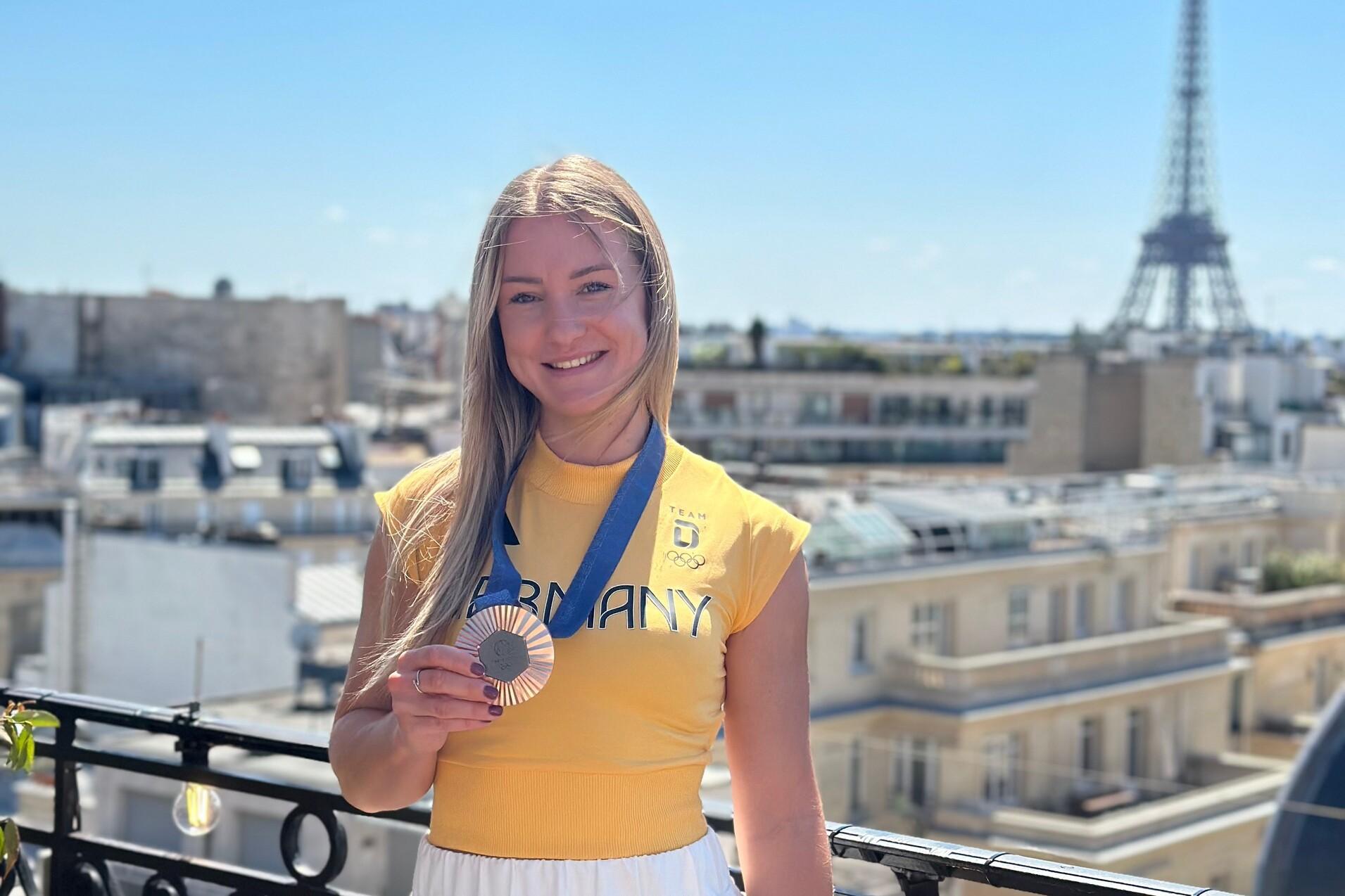 Junge Athletin in gelbem Shirt zeigt stolz ihre Medaille vor dem Eiffelturm und einem klaren blauen Himmel.