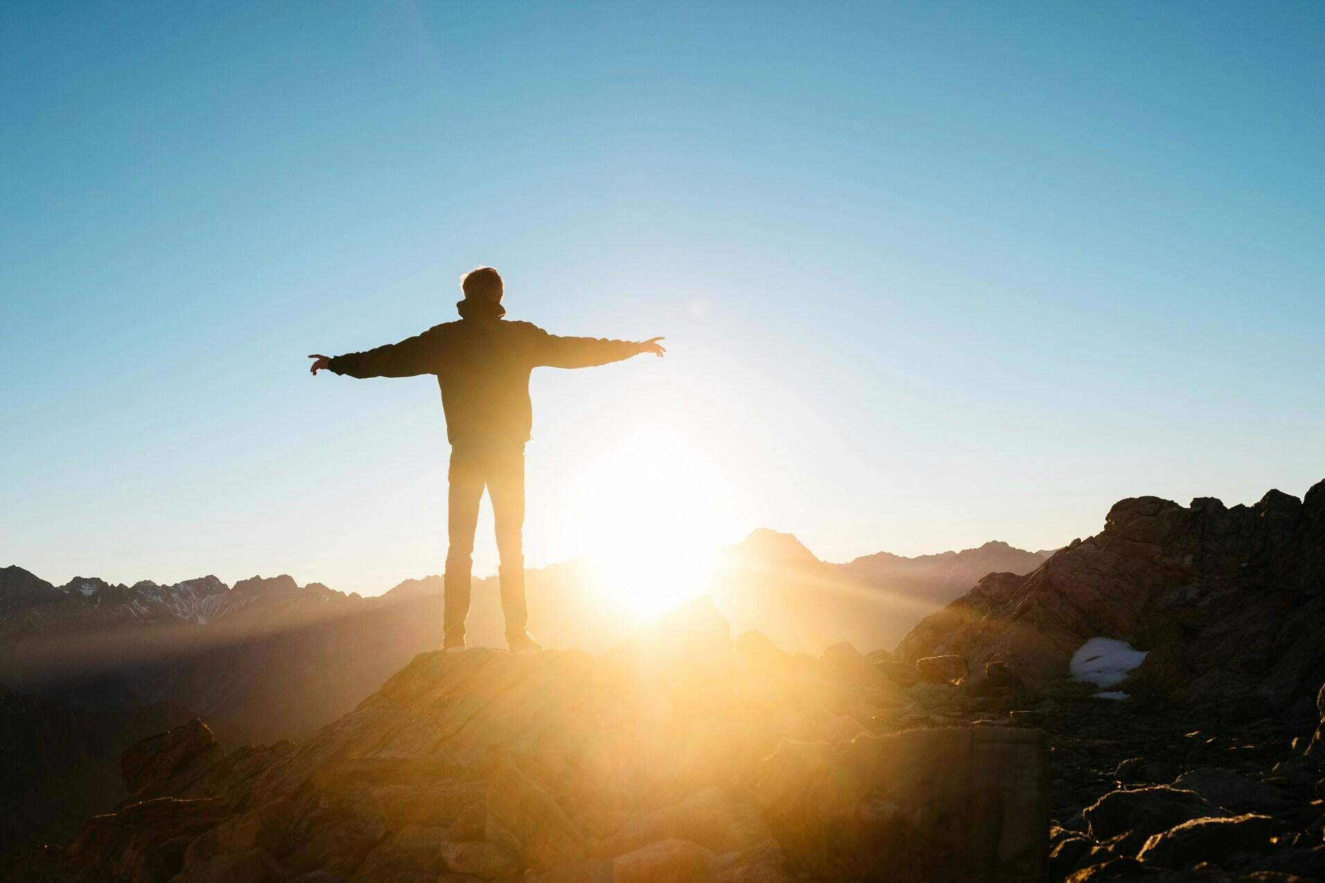 Silhouette eines stehenden Menschen mit ausgestreckten Armen auf einem Felsen bei Sonnenaufgang in den Bergen.