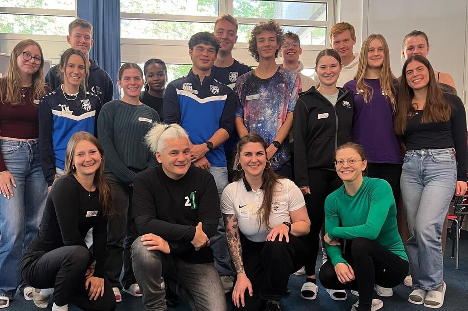 Gruppenbild von 20 Personen mit unterschiedlichen Outfits in einem hellen Raum, einige lächeln, andere posieren neutral.