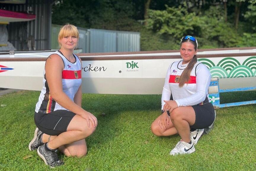 Zwei Ruderinnen knien auf gr&uuml;nem Gras neben einem Ruderboot, umgeben von bunten Fahnen und B&auml;umen.