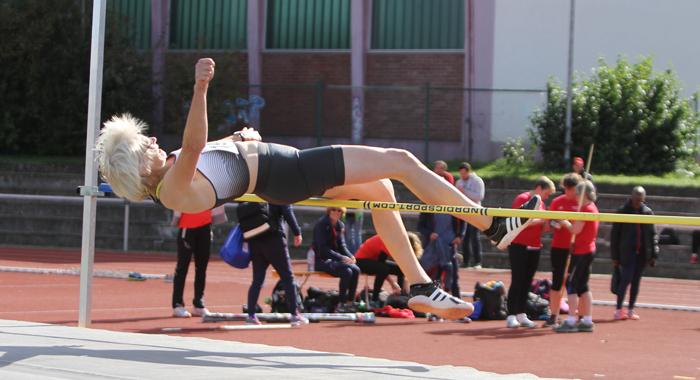 Sportlerin springt &uuml;ber eine Hochsprunglatte, w&auml;hrend Zuschauer im Hintergrund ihre Technik beobachten.