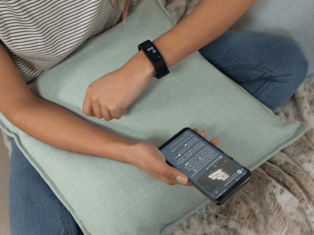 Junge Frau mit Fitness-Armband schaut auf ihr Smartphone, w&auml;hrend sie auf einem Kissen sitzt.