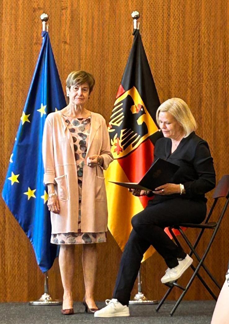 Zwei Frauen stehen auf einer B&uuml;hne, umgeben von Europaflagge und Bundesflagge, eine h&auml;lt eine Mappe.