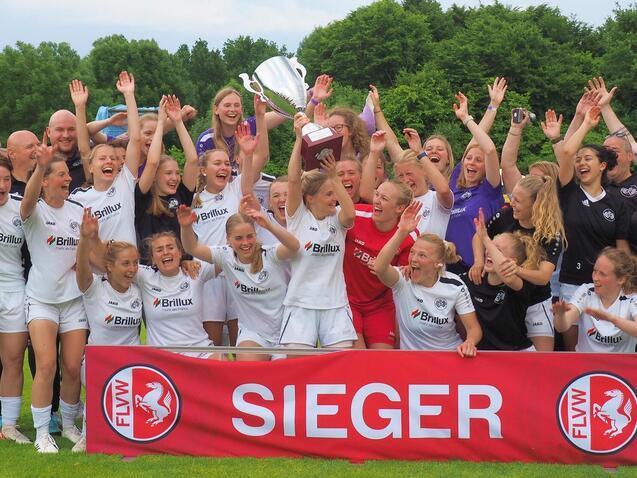 Mannschaft feiert mit Pokal und Banner "SIEGER" auf dem Fu&szlig;ballplatz, umgeben von jubelnden Spielerinnen und Unterst&uuml;tzern.