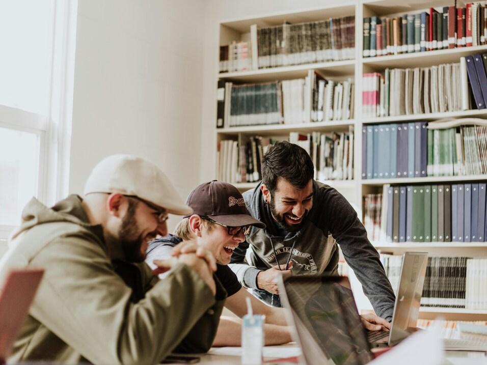 F&uuml;nf Personen sitzen an einem Tisch, lachen und schauen auf einen Laptop vor Regalen mit B&uuml;cherordnern.