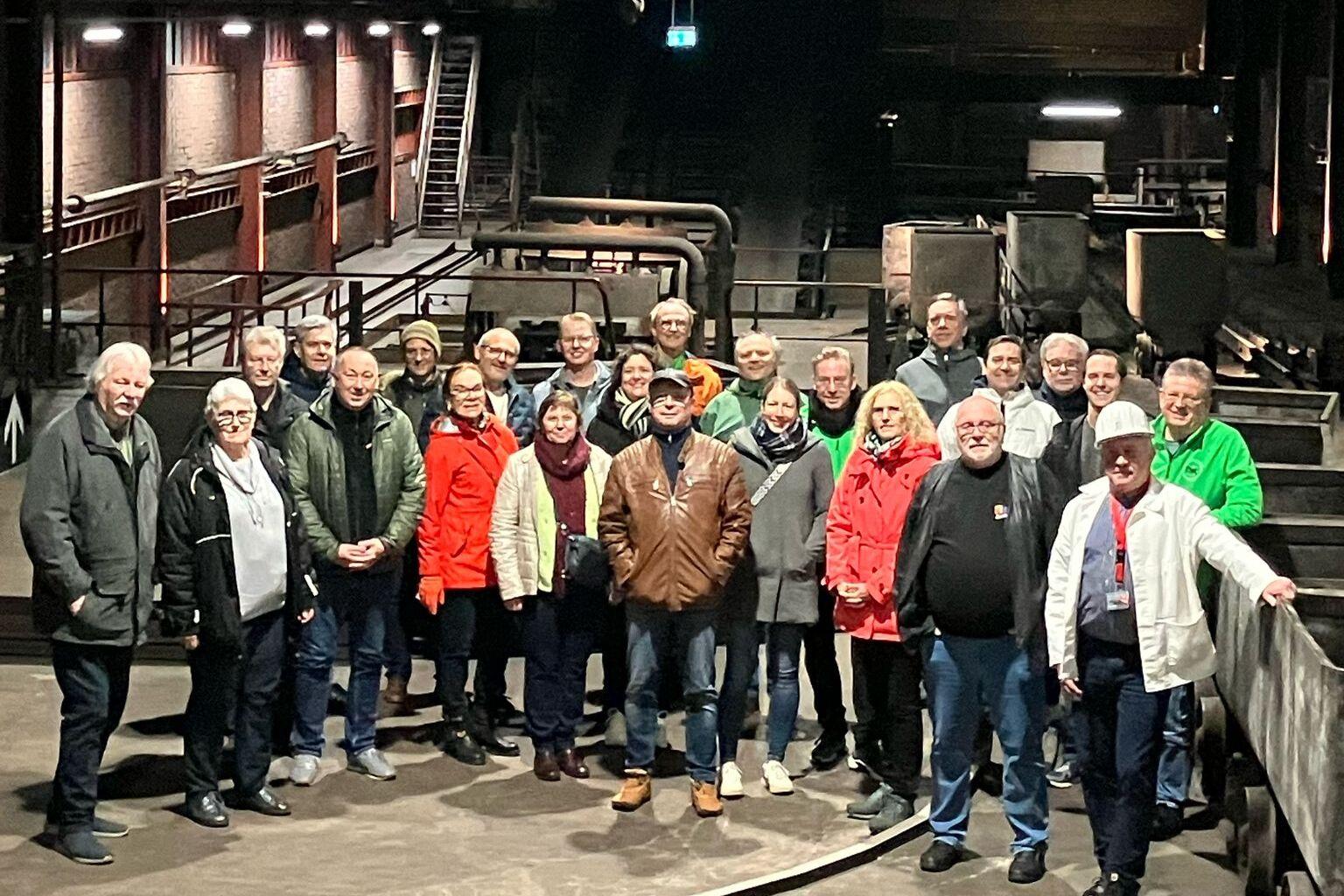 Gruppe von 30 Personen in einer Industriehalle, umgeben von Rohren und Maschinen, bei einer Besichtigung.