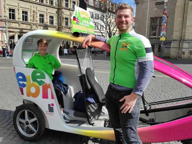 Zwei Personen in gr&uuml;nen Shirts stehen neben einem bunten Pedelac in einer Stadt, im Hintergrund historische Geb&auml;ude.