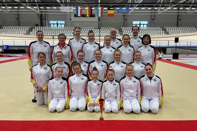 Gruppe von Sportlerinnen in einheitlichen Trainingsanz&uuml;gen mit Pokal auf einer Turnmatte in einer Sporthalle.