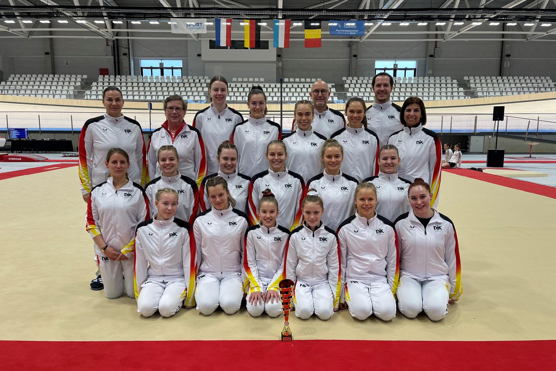 Gruppe von Sportlerinnen in einheitlichen Trainingsanz&uuml;gen mit Pokal auf einer Turnmatte in einer Sporthalle.