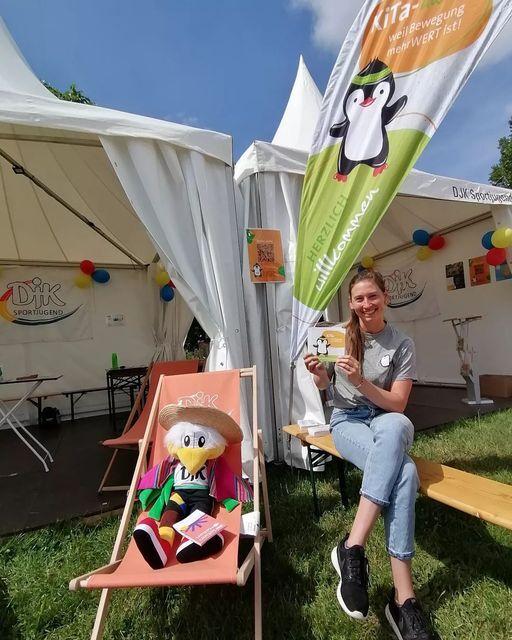 Eine Frau sitzt l&auml;chelnd auf einer Bank, h&auml;lt eine Flagge und posiert neben einem Pl&uuml;schtier auf einem Liegestuhl.