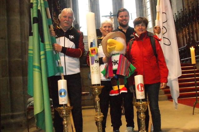 Vier Personen in Kirchenumgebung halten Fahnen und eine Maskottchenfigur zwischen zwei goldenen Kerzenhaltern.