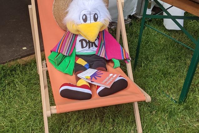 Pl&uuml;schfigur eines bunten Vogels in einem liegenden Stuhl mit einem Sonnenhut und bunten Kleidungsst&uuml;cken.